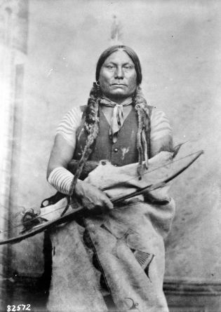 The sombre chief stares into the camera, upper parts plainly dressed, a single eagle feather in his hair, twin braids.  He holds a bow in his right hand and has a blanket around his waist.