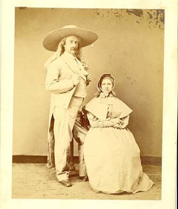 Custer, long hair past his shoulders, thumbs in lapels, is in a pale suit,  with a jacket whose tails nearly brush the floor, and wearing the biggest brimmed hat you ever saw.  His bonneted sister sits demurely at his left.