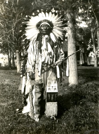 The chief is in full regalia, including full feather headdress and ceremonial pipe, trees in the near background